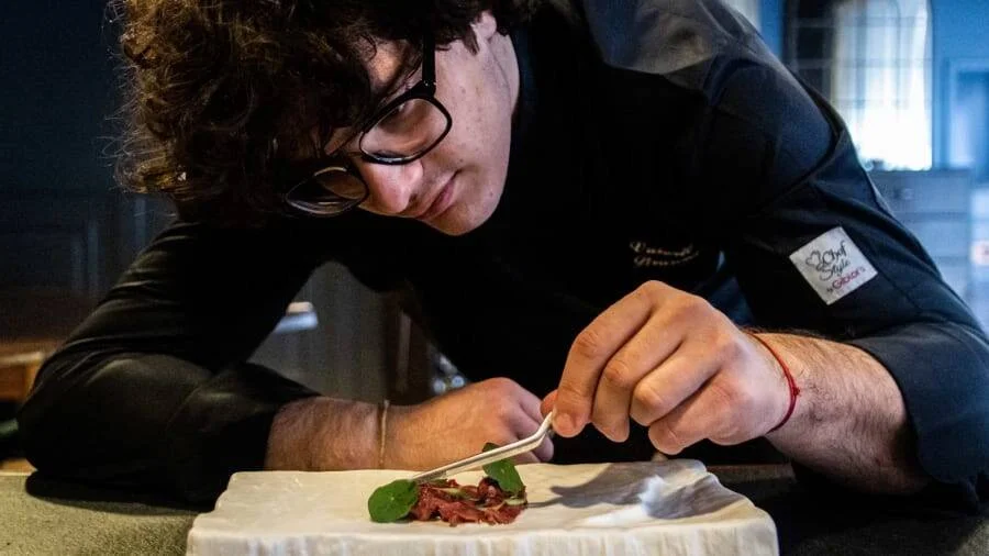 Joven chef con uniforme negro coloca con pinzas una hoja verde sobre un plato de carne con precisión.