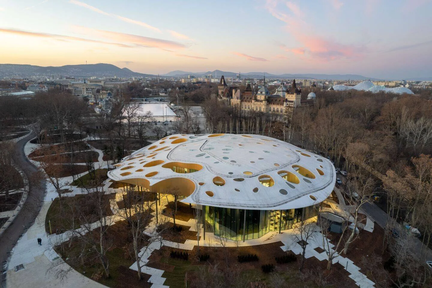 Vista aérea al atardecer de un edificio contemporáneo de techo ovalado con aberturas circulares, rodeado de árboles en un parque de Budapest.