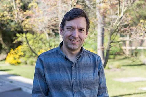 Hombre de mediana edad con camisa gris a cuadros sonríe en un entorno verde al aire libre.