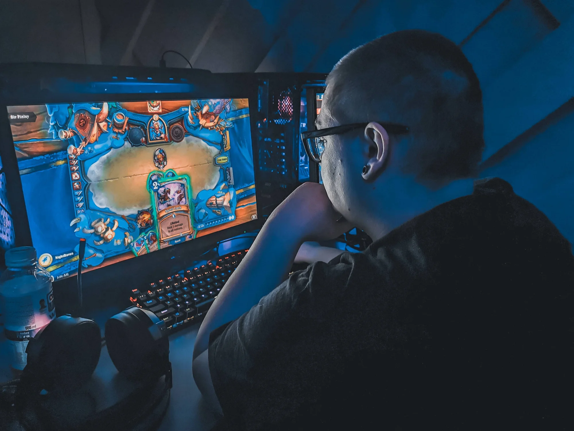 Joven con gafas juega un videojuego de cartas en su computadora de escritorio en una habitación oscura iluminada por la pantalla.
