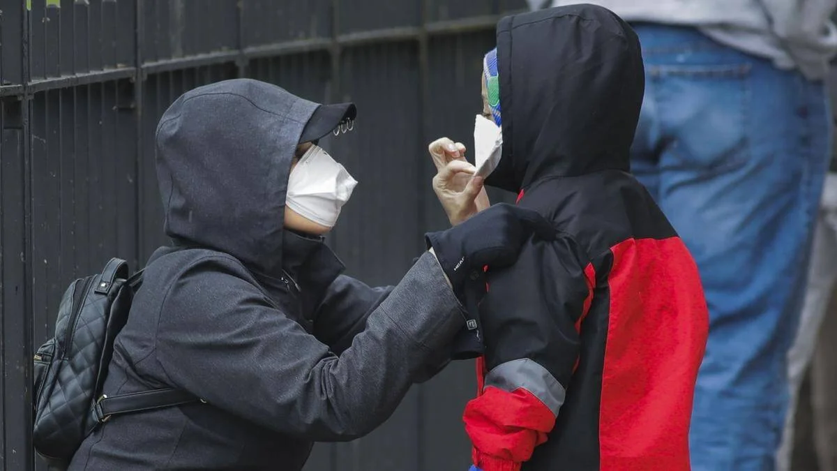 Una persona con tapabocas ajusta la mascarilla a un niño con capucha en la vía pública.