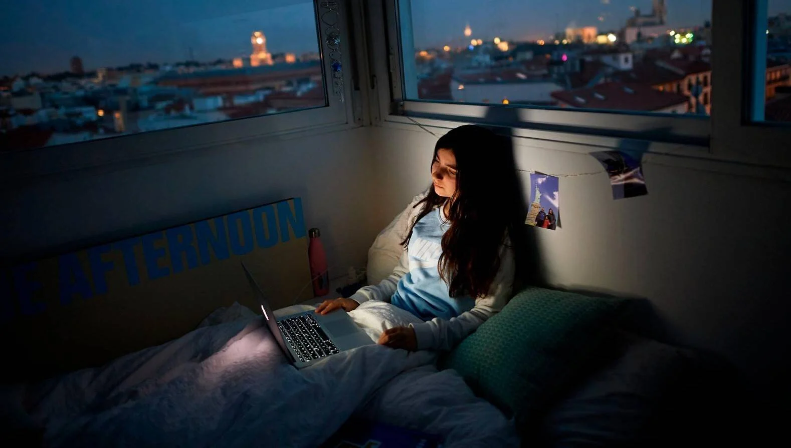 Adolescente acostada en su cama usando un portátil en la oscuridad, iluminada por la pantalla, con vista nocturna de ciudad al fondo.