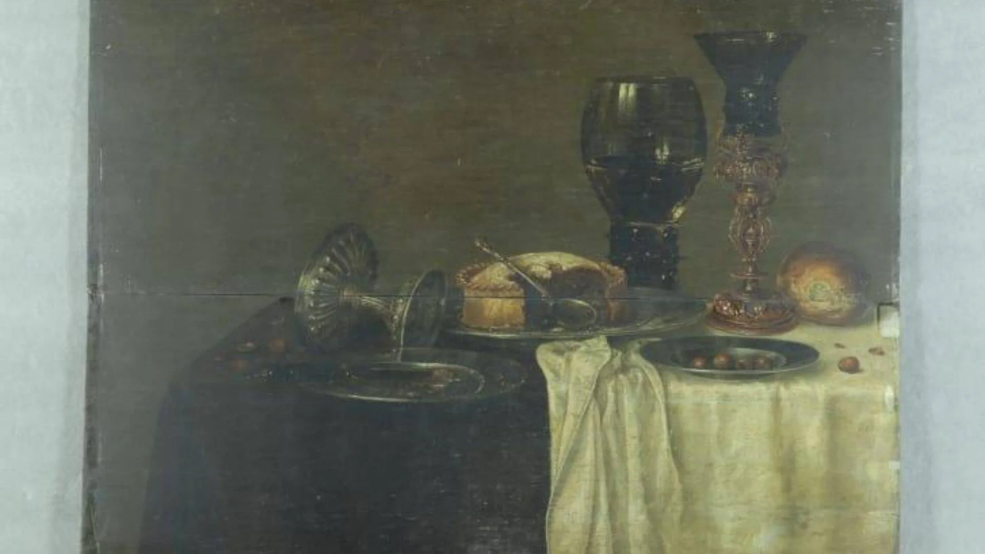 Óleo de estilo flamenco con naturaleza muerta: copas de cristal, copa ornamental, pastel en bandeja, frutos y mantel blanco sobre mesa.