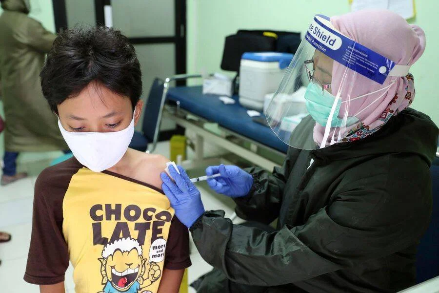 Una trabajadora de salud con escudo facial y guantes aplica una vacuna en el brazo de un niño con mascarilla.