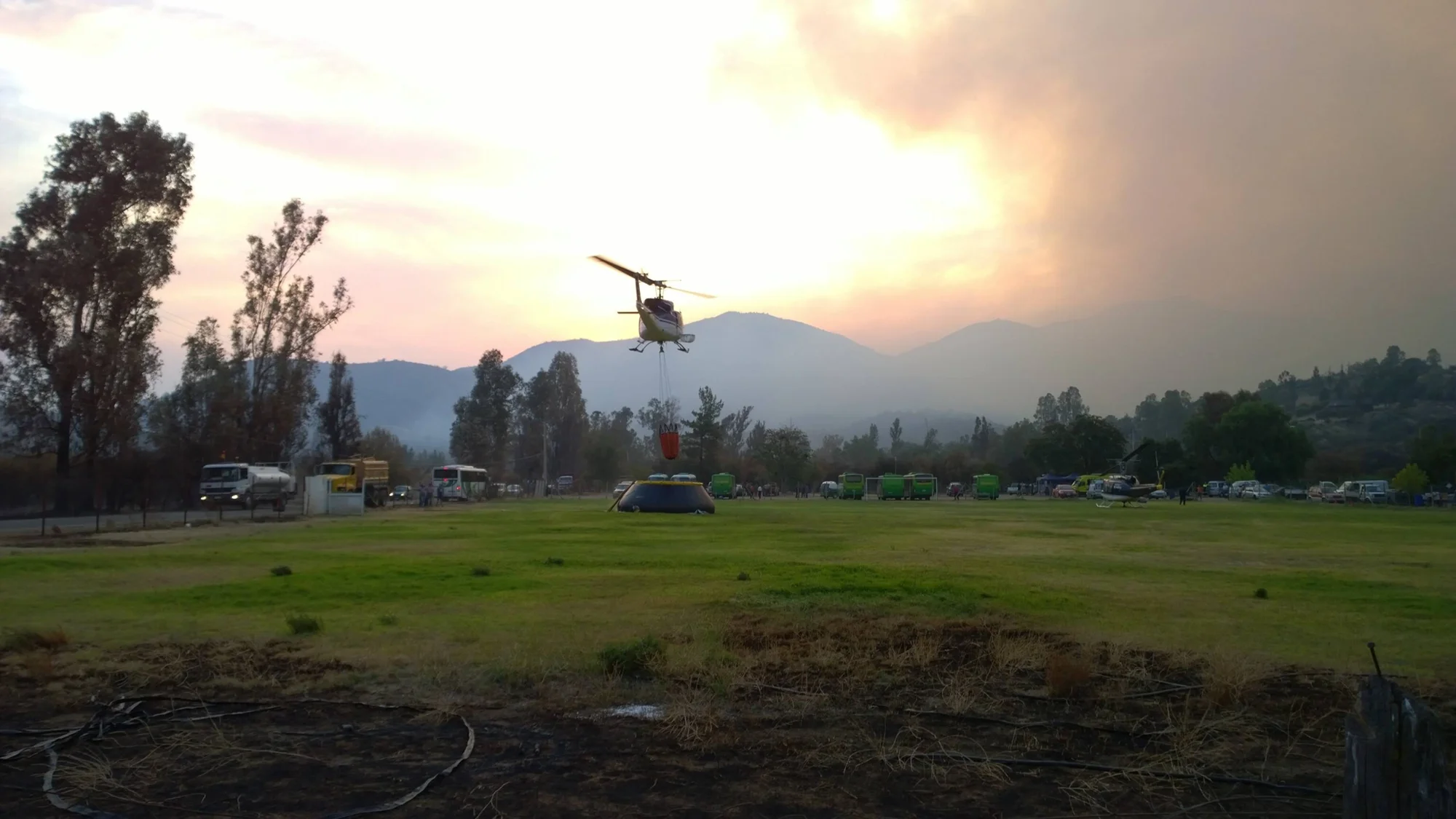 Helicóptero con cubeta cargando agua en campo de operaciones rodeado de vehículos de emergencia, con humo y montañas al fondo.