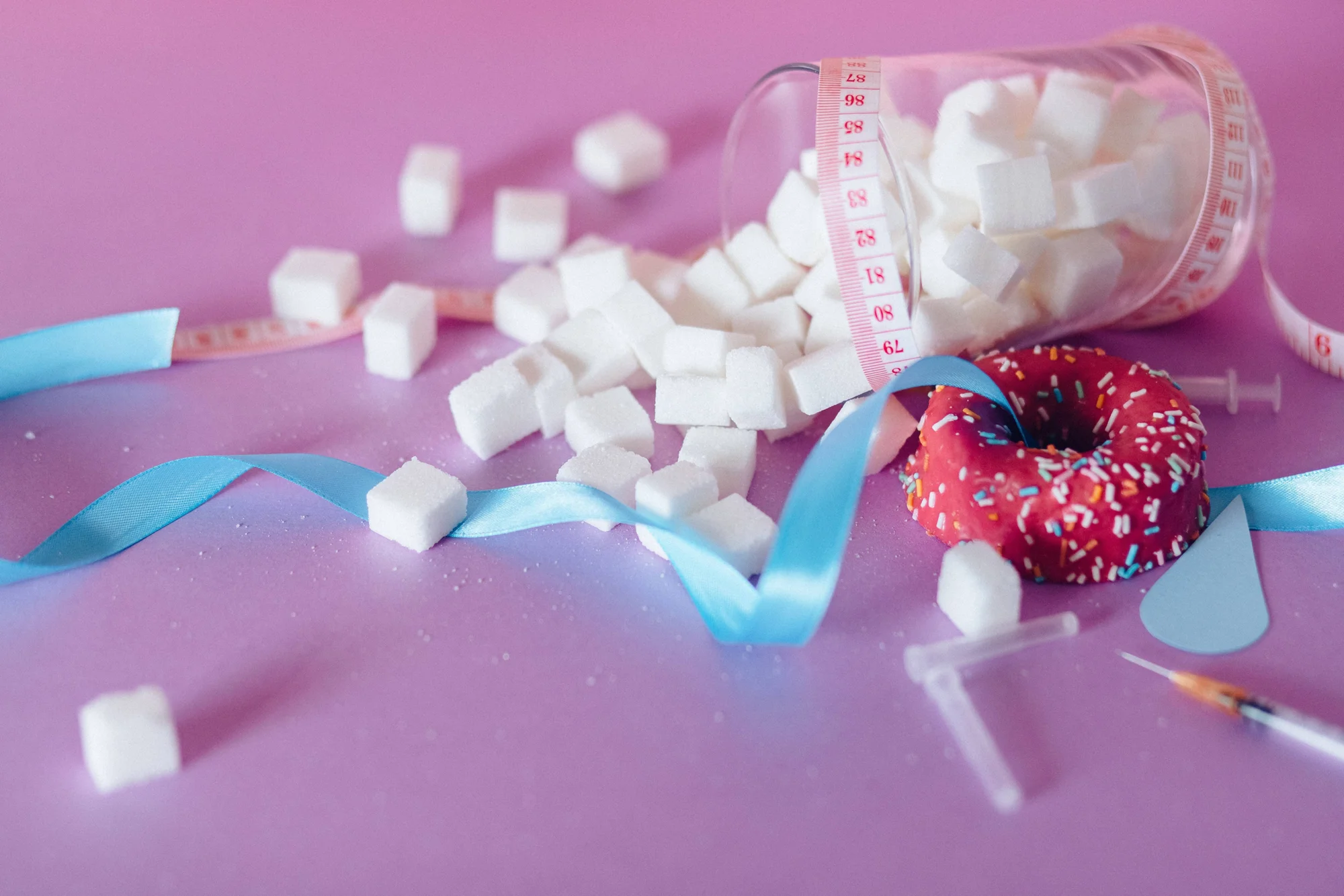 Frasco volcado con terrones de azúcar, una dona con glaseado rojo, cinta métrica y jeringa, simbolizando el consumo excesivo de azúcar y la diabetes.