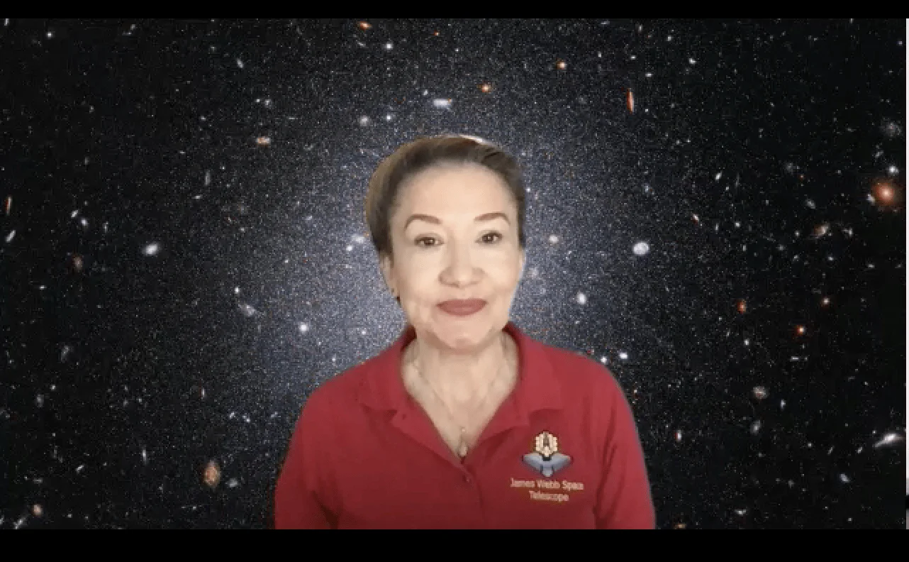 Mujer con camiseta del James Webb Space Telescope habla a cámara con fondo de imagen del universo profundo.