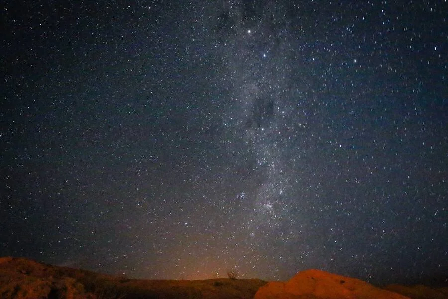 La Vía Láctea se extiende sobre un cielo estrellado con colinas rojizas en el horizonte bajo luz nocturna.