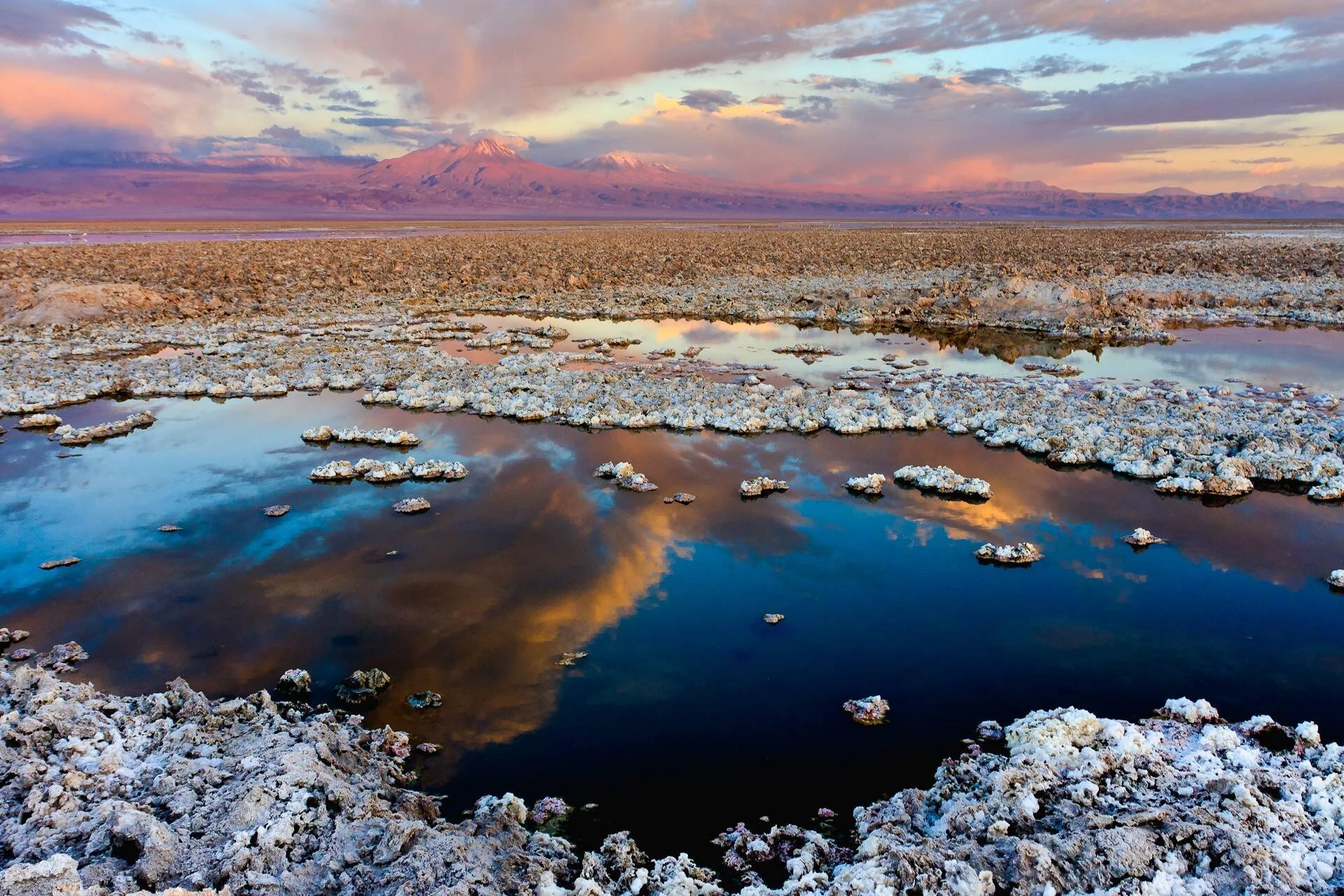 Salar con cristales de sal y pozas de agua que reflejan un cielo rojizo al atardecer, con un volcán nevado al fondo.