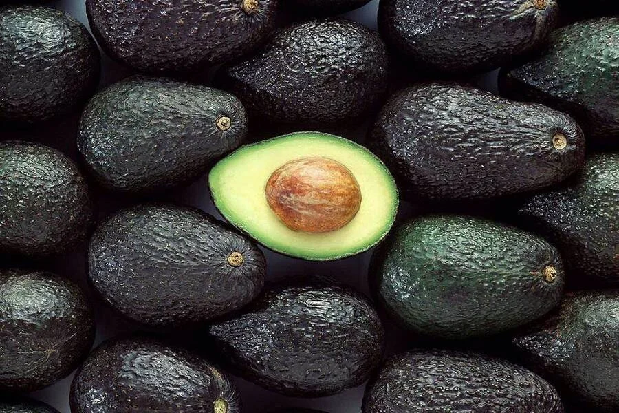 Varios aguacates maduros de cáscara oscura junto a una mitad abierta que muestra su pulpa verde y hueso.