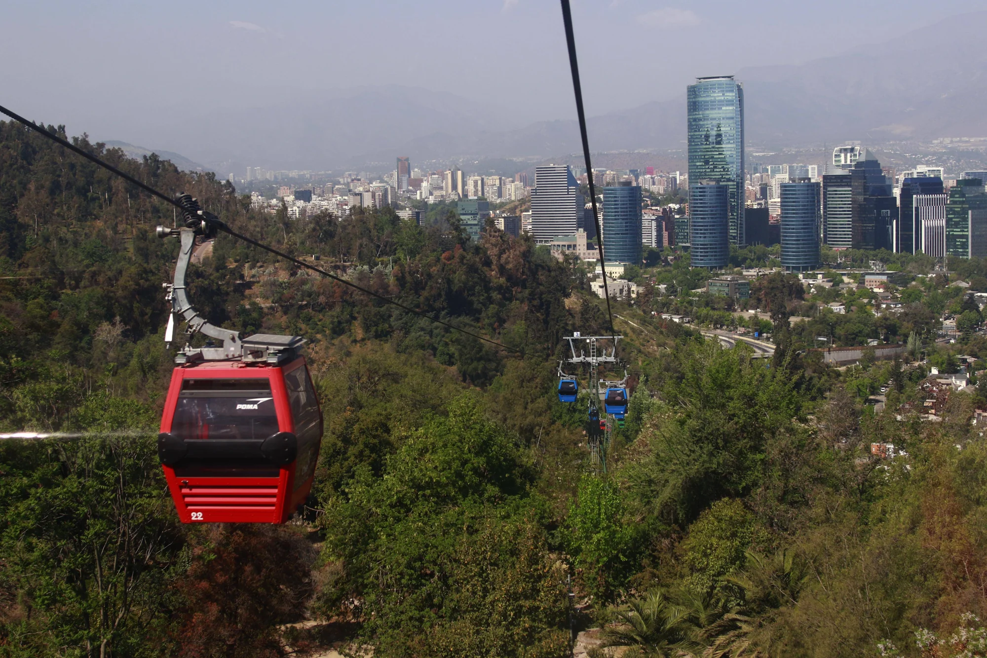 Cabinas de teleférico roja y azul descienden sobre un cerro arbolado con el skyline de Santiago de Chile al fondo.