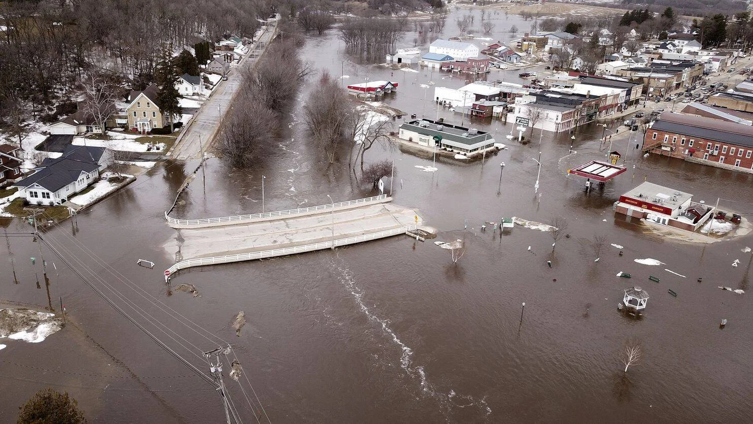 Vista aérea de una localidad completamente inundada, con calles, puente y edificios comerciales cubiertos por agua marrón.