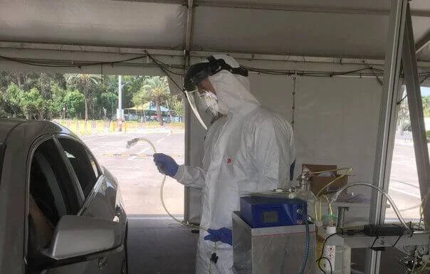 Profesional de salud con traje protector blanco y escudo facial atiende a un conductor en un puesto de testeo de COVID-19 bajo carpa.