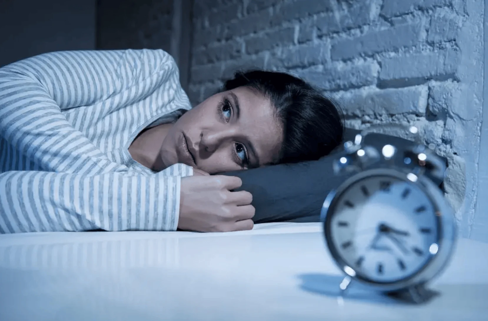 Mujer acostada en la cama con los ojos abiertos mirando un despertador, incapaz de dormir de noche.