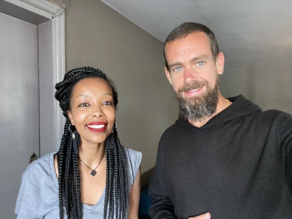 Una mujer con trenzas largas y labial rojo junto a un hombre con barba y sudadera negra, sonriendo a la cámara.