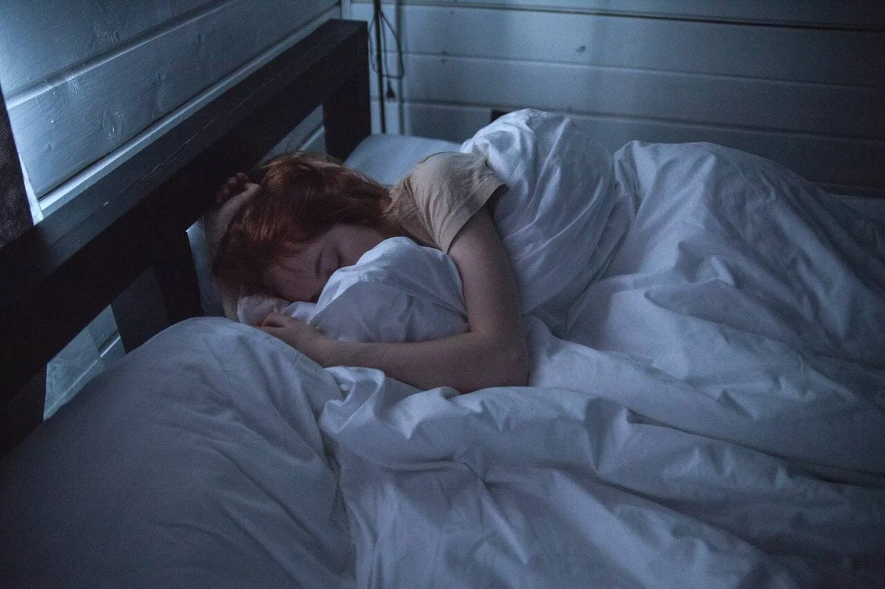 Mujer pelirroja dormida en una cama con sábanas blancas en una habitación oscura.