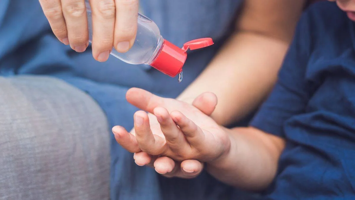 Una persona adulta vierte gel desinfectante de un frasco con tapa roja sobre la mano abierta de un niño pequeño.