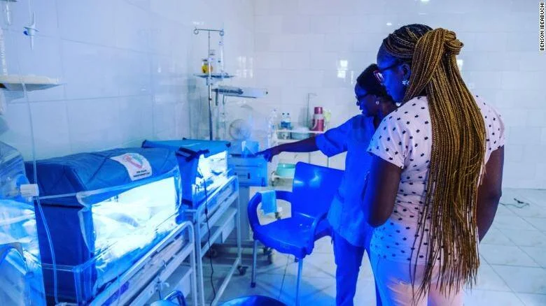 Una enfermera y una mujer observan a un recién nacido en una incubadora con luz azul en una sala hospitalaria.