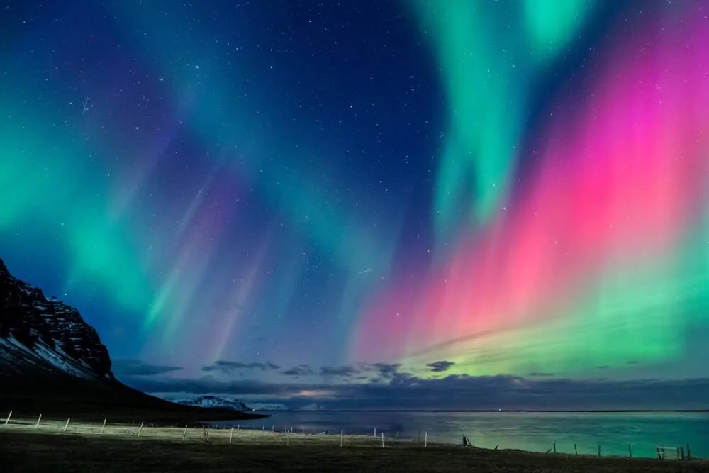Espectacular aurora boreal con bandas de luz verde y rosa iluminando el cielo nocturno sobre una costa ártica montañosa.