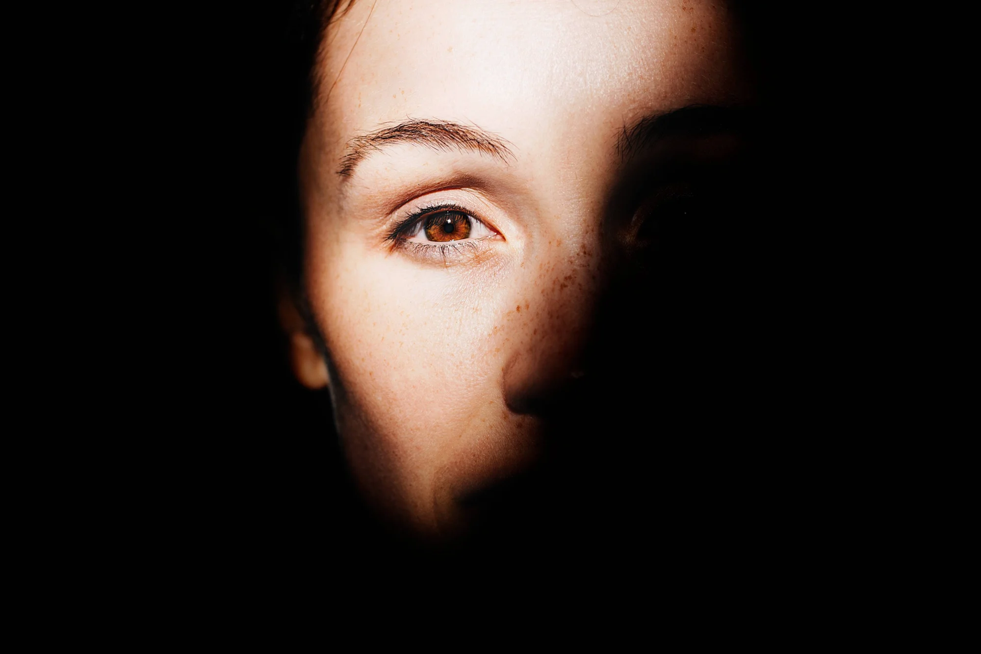 Mitad del rostro de una mujer con ojo marrón iluminado, parcialmente cubierto por sombras sobre fondo negro.