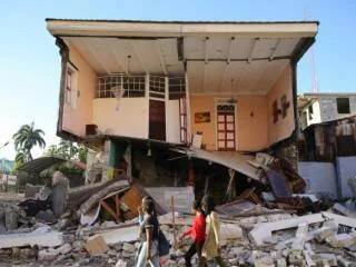Personas caminan frente a un edificio con su planta baja destruida por un sismo, con escombros en el suelo.