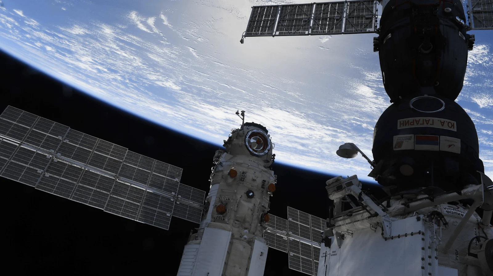 Módulos y paneles solares de la ISS con inscripción en ruso, orbitando sobre la Tierra con su atmósfera visible al fondo.