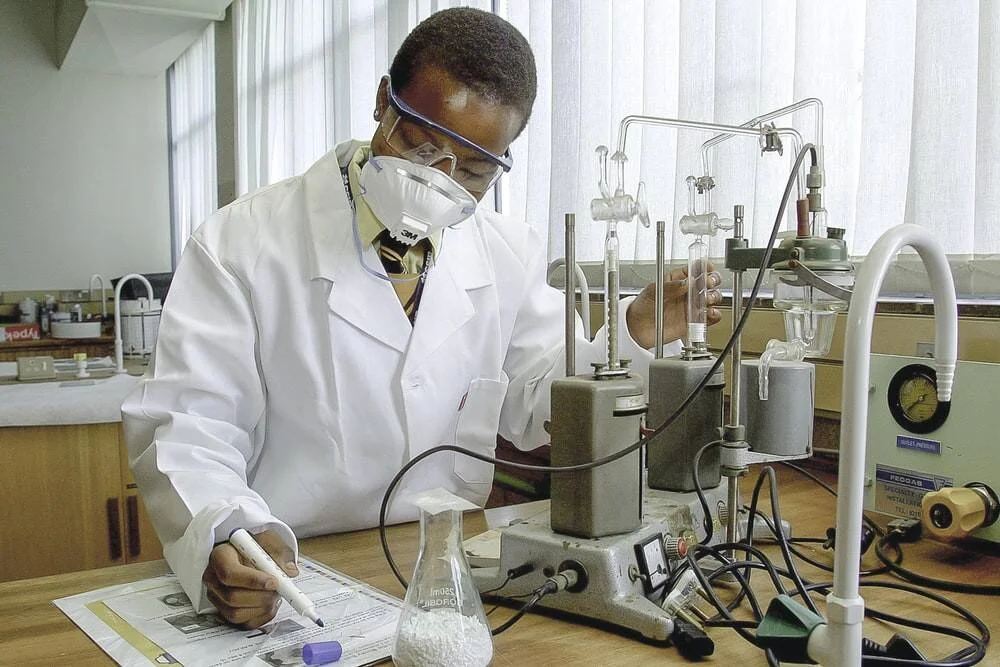 Investigador con bata blanca, mascarilla y gafas de seguridad toma notas junto a equipos de laboratorio y un matraz Erlenmeyer.