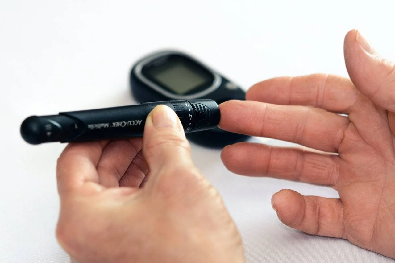 Persona aplicando una lanceta en el dedo para medir el nivel de glucosa en sangre, con un glucómetro al fondo.