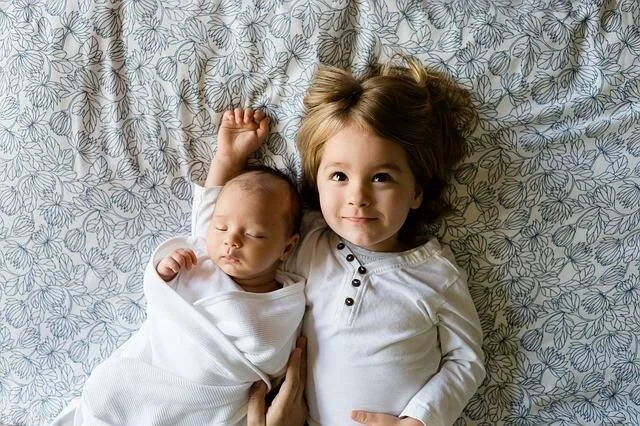 Un bebé recién nacido dormido envuelto en blanco junto a un niño pequeño sonriente, tumbados sobre una sábana floral.