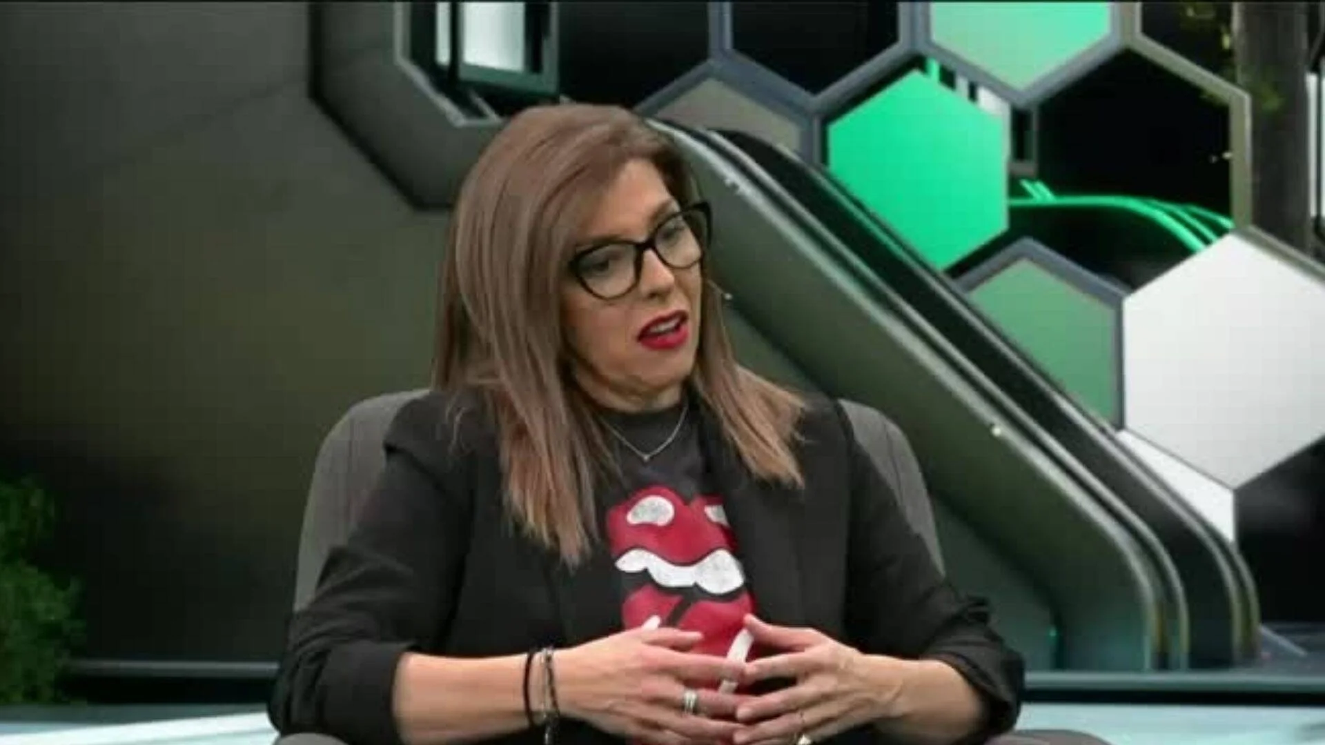 Mujer con gafas, remera de los Rolling Stones y blazer negro sentada en un estudio de televisión con decorado hexagonal verde y negro.