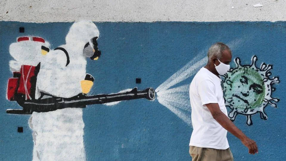 Un hombre con mascarilla camina frente a un mural que representa a un trabajador con traje de protección fumigando un coronavirus.