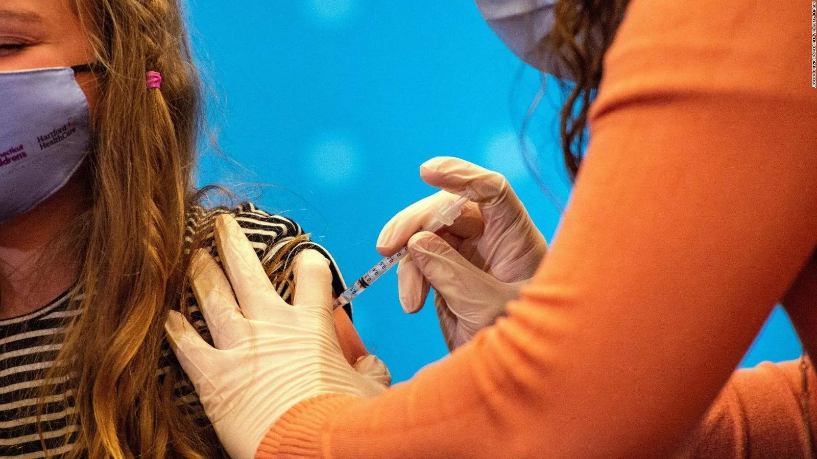 Un profesional de salud con guantes aplica una inyección en el brazo de una niña que lleva mascarilla quirúrgica.