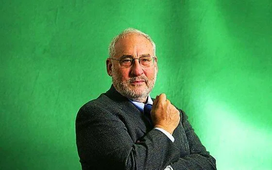 Hombre de cabello canoso y barba gris, con saco gris, posa con los brazos cruzados frente a un fondo verde.
