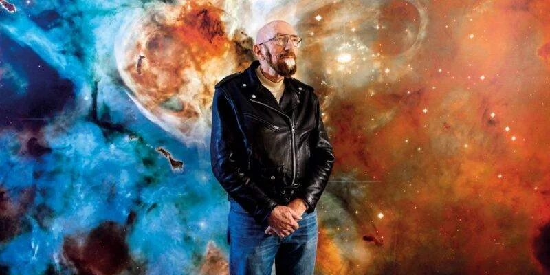 Hombre calvo con barba y chaqueta de cuero negro posa frente a un gran mural de nebulosas coloridas del cosmos.