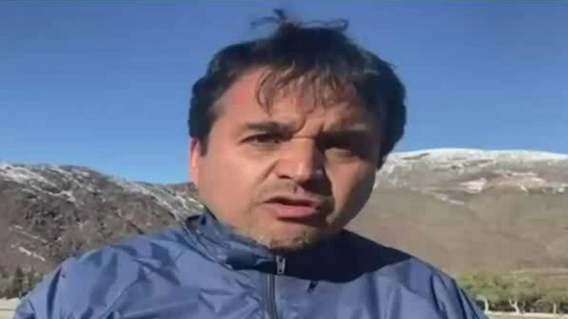 Hombre de cabello oscuro con campera azul habla a cámara con montañas nevadas al fondo.