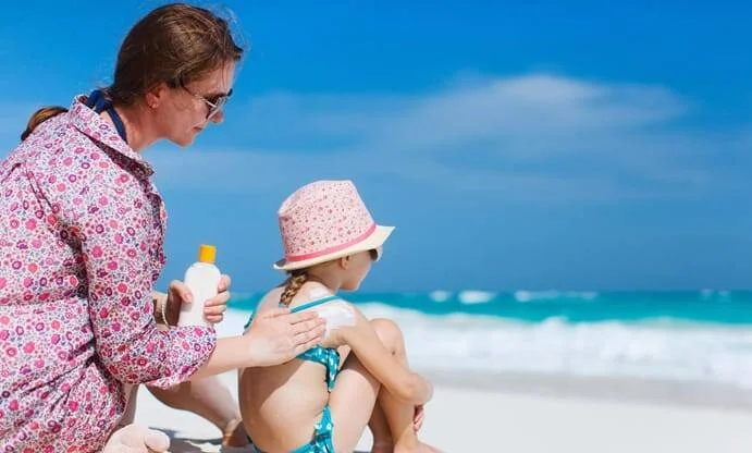 Una persona aplica protector solar en la espalda de una niña con sombrero rosa sentada en la playa.
