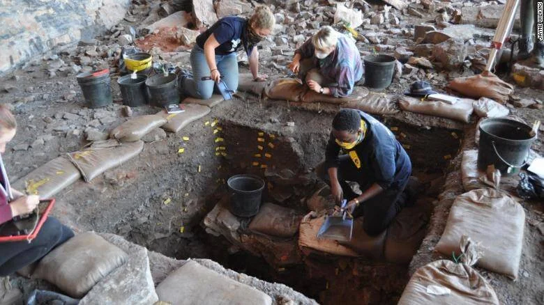 Equipo de arqueólogos excava cuidadosamente una cueva rocosa con herramientas manuales y marcadores amarillos en el suelo.