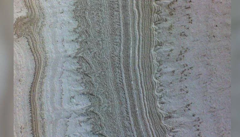 Vista aérea de capas de hielo y polvo en la superficie marciana, con bandas onduladas en tonos gris verdoso y beige.