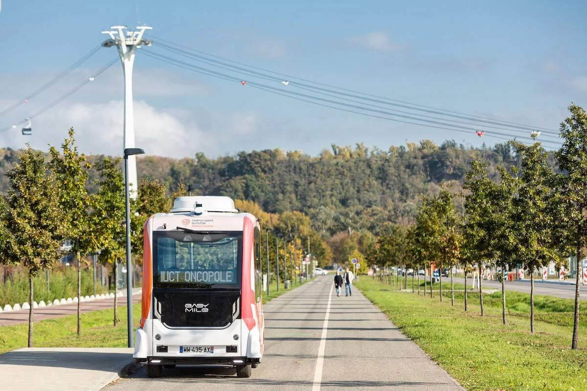 Vehículo autónomo EasyMile con destino IUCT Oncopole circula por una avenida arbolada del Campus Santé du Futur.
