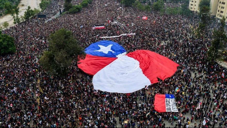 Vista aérea de una masiva manifestación en Chile con una enorme bandera chilena desplegada en medio de la multitud.
