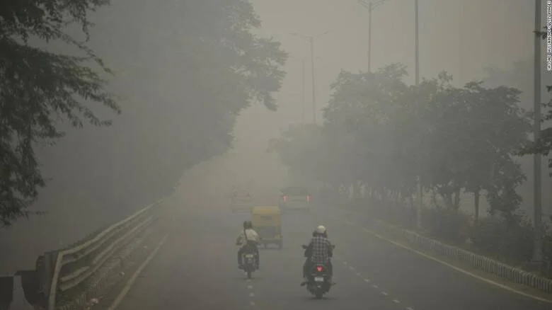 Motociclistas circulan por una avenida arbolada envuelta en densa neblina de contaminación con visibilidad muy reducida.