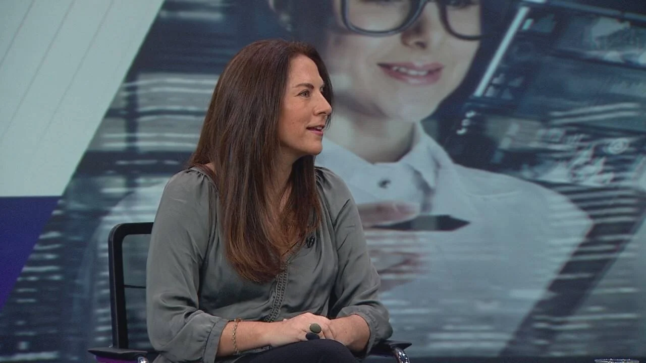 Mujer de cabello castaño largo sentada en estudio de televisión frente a una pantalla gráfica de fondo.