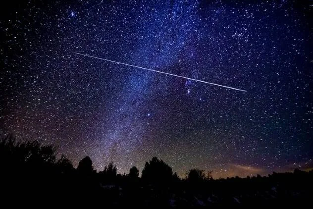 Un meteoro deja un rastro luminoso sobre un cielo estrellado con la Vía Láctea visible, sobre la silueta de árboles.