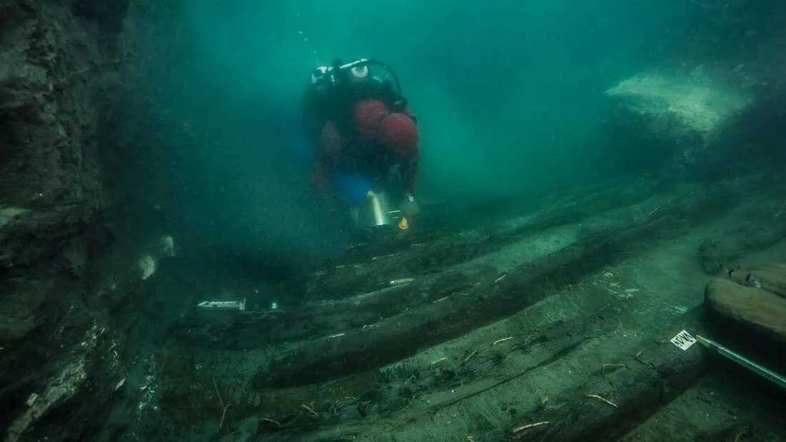 Un buzo con traje rojo inspecciona los restos de madera de un naufragio antiguo en el fondo marino.