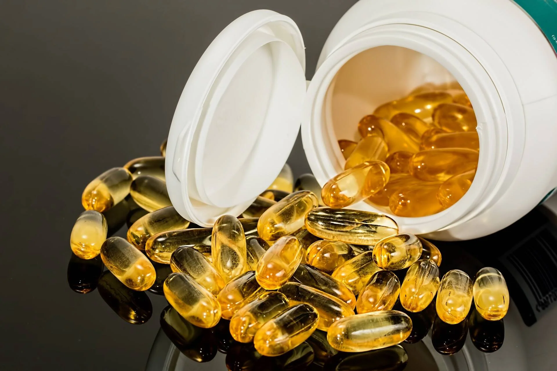 Cápsulas blandas de color ámbar, posiblemente omega-3 o aceite de pescado, derramadas desde un frasco blanco abierto sobre una superficie reflectante.
