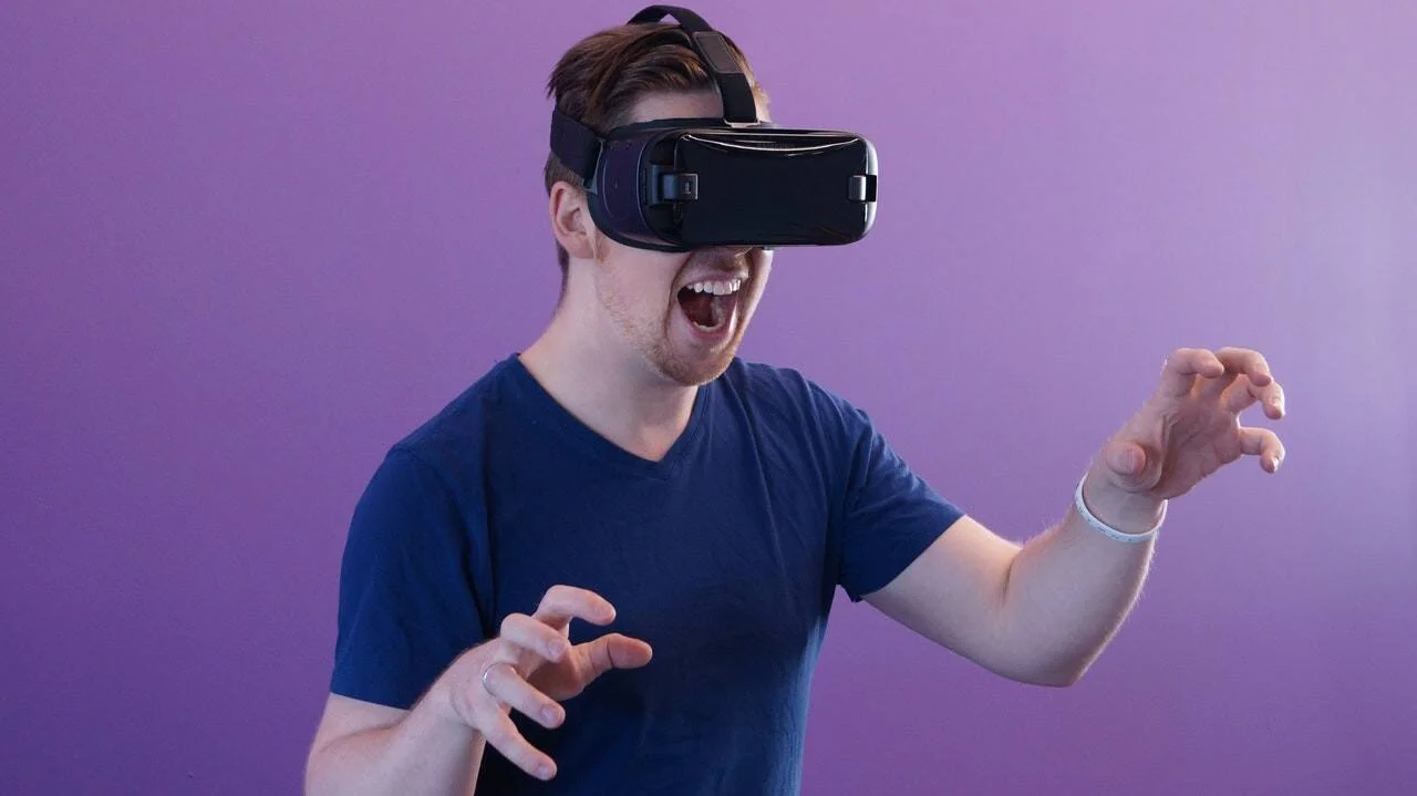Hombre joven con casco de realidad virtual reacciona con entusiasmo y las manos extendidas ante una experiencia inmersiva.