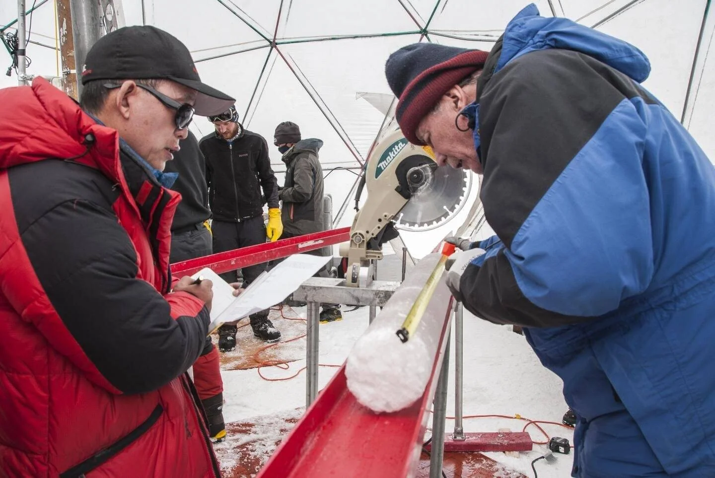 Investigadores examinan y cortan un testigo de hielo con sierra circular en una carpa de campo polar.