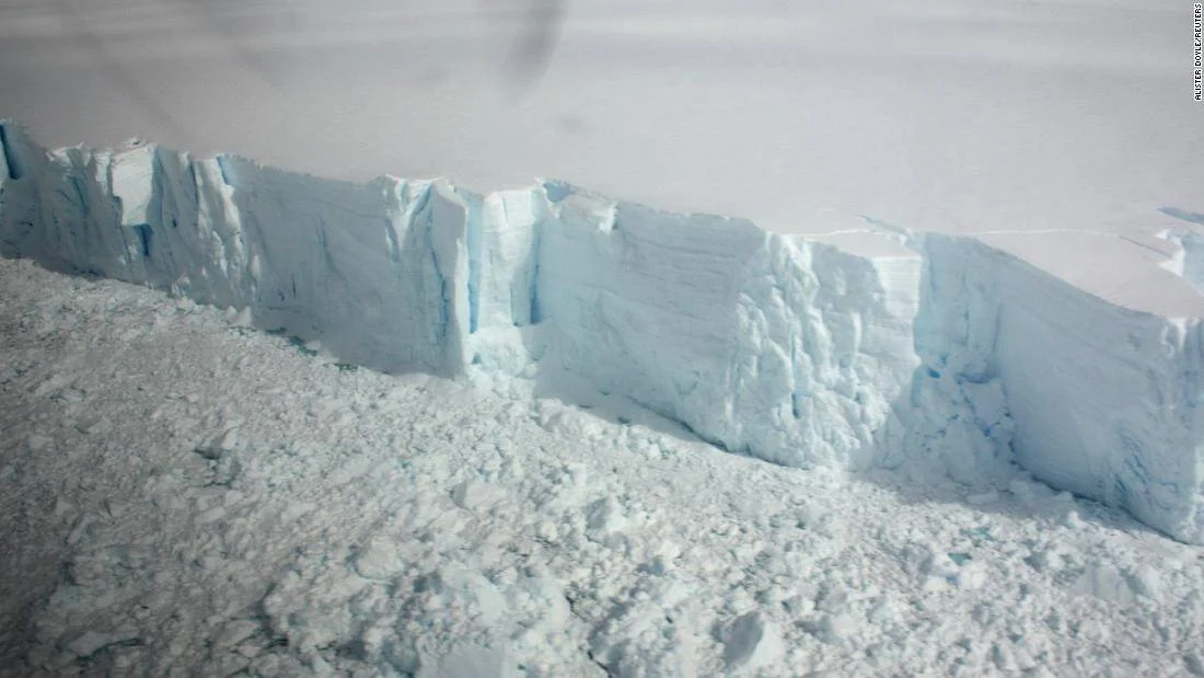 Vista aérea del borde vertical de un glaciar antártico con paredes de hielo azul y escombros de hielo en la base.