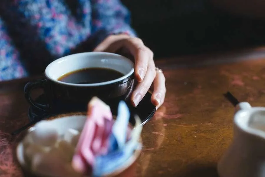 Una persona sostiene una taza oscura con café negro sobre una mesa de madera, con servilletas y azucarera al fondo.