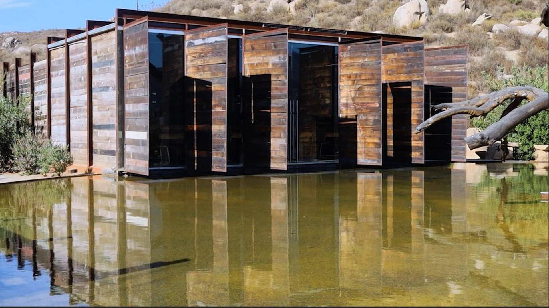 Construcción de madera rústica y vidrio negro junto a un estanque que refleja su imagen, rodeada de paisaje árido y rocas.